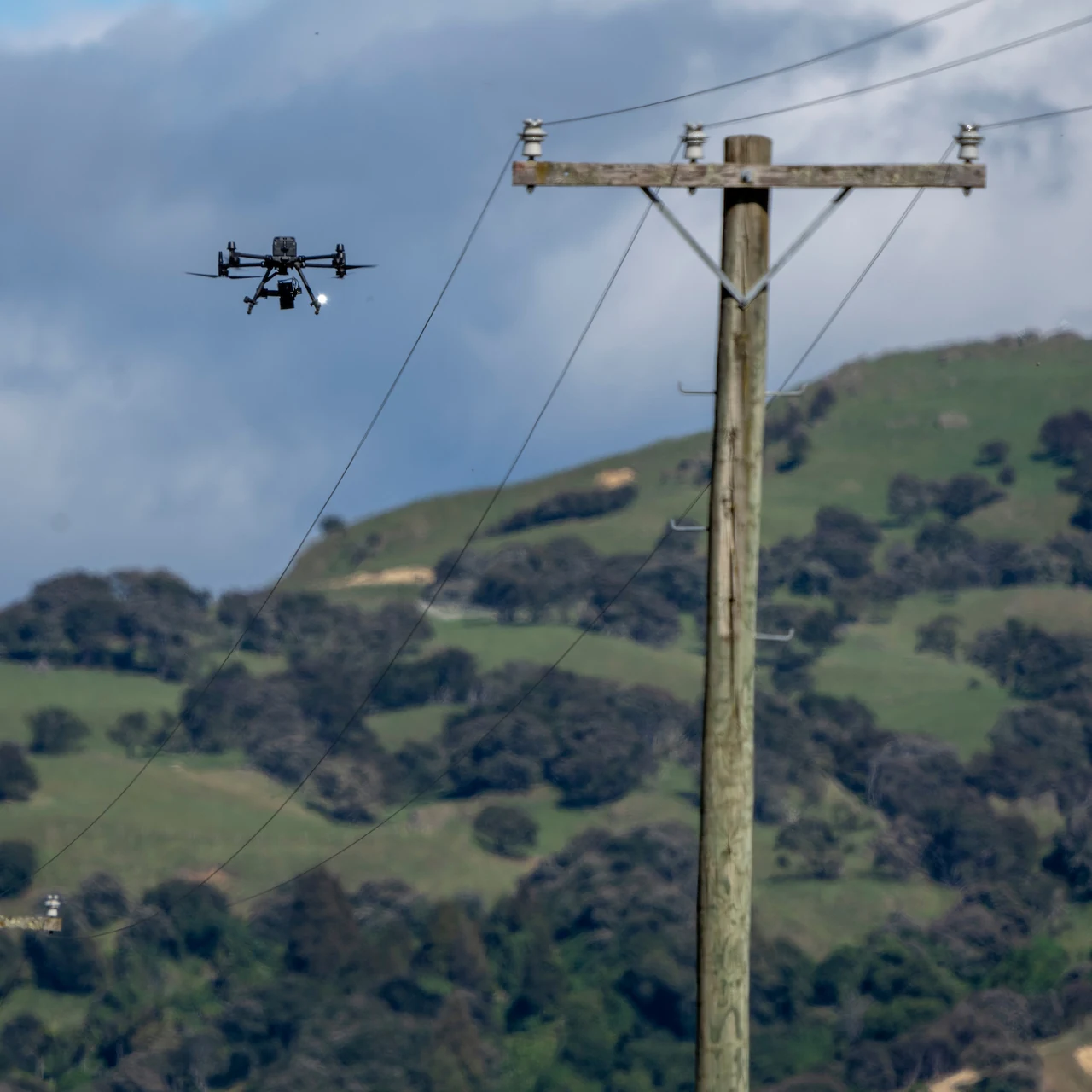 An inspection drone flying alongside a wooden crossarm pole and its conductors above New Zealand hill country.