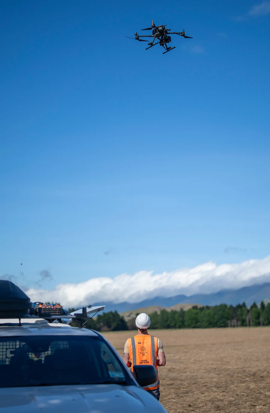 A drone operator in high-visibility gear flying an inspection drone beside an equipped field vehicle.