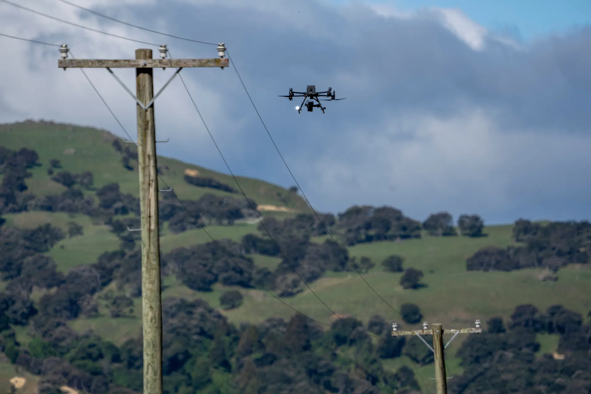 An inspection drone flying alongside the conductors of a rural overhead distribution line in New Zealand hill country.